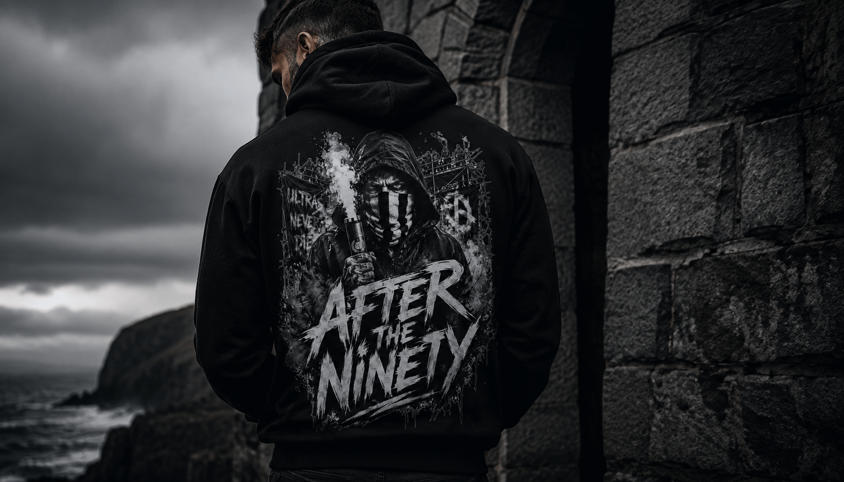 A person viewed from behind wearing a black hoodie with a large white graphic print on the back that reads 'AFTER THE NINETY' and shows a stone wall and dark ocean in the background.