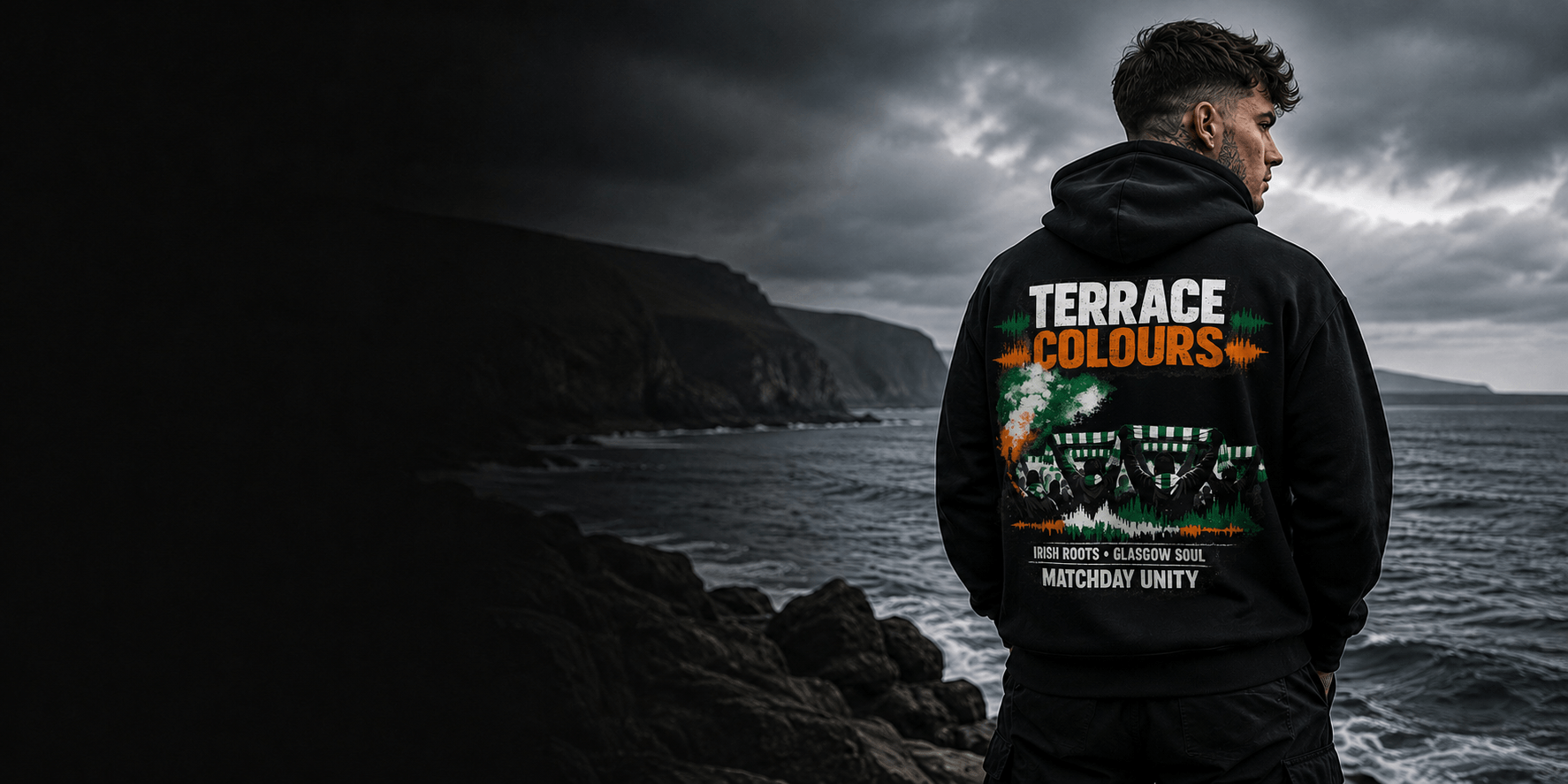 Model wearing a black Celtic Wolfwear Terrace Colours graphic hoodie beside a rugged coastline