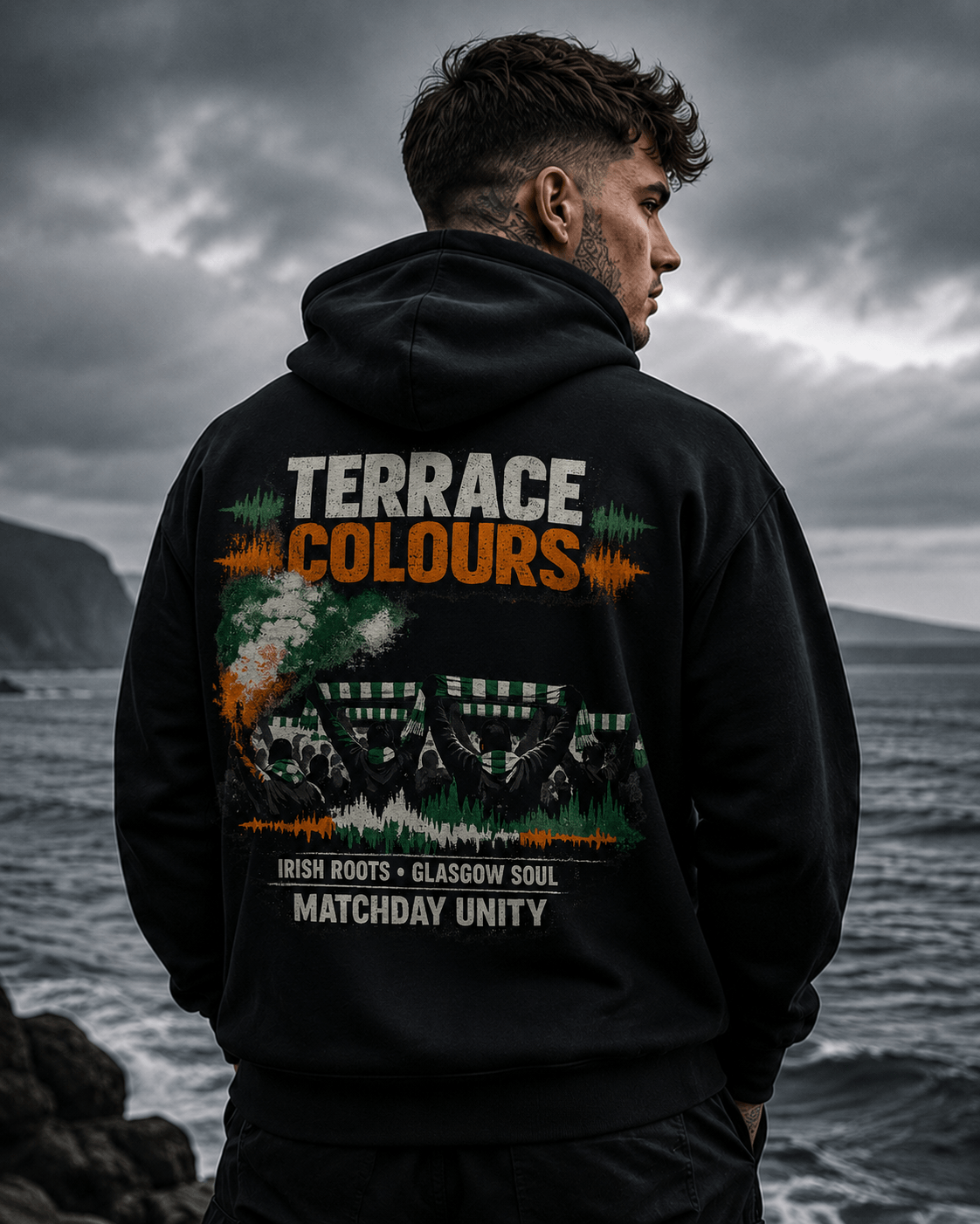 Model wearing a black Celtic Wolfwear Terrace Colours graphic hoodie beside a rugged coastline